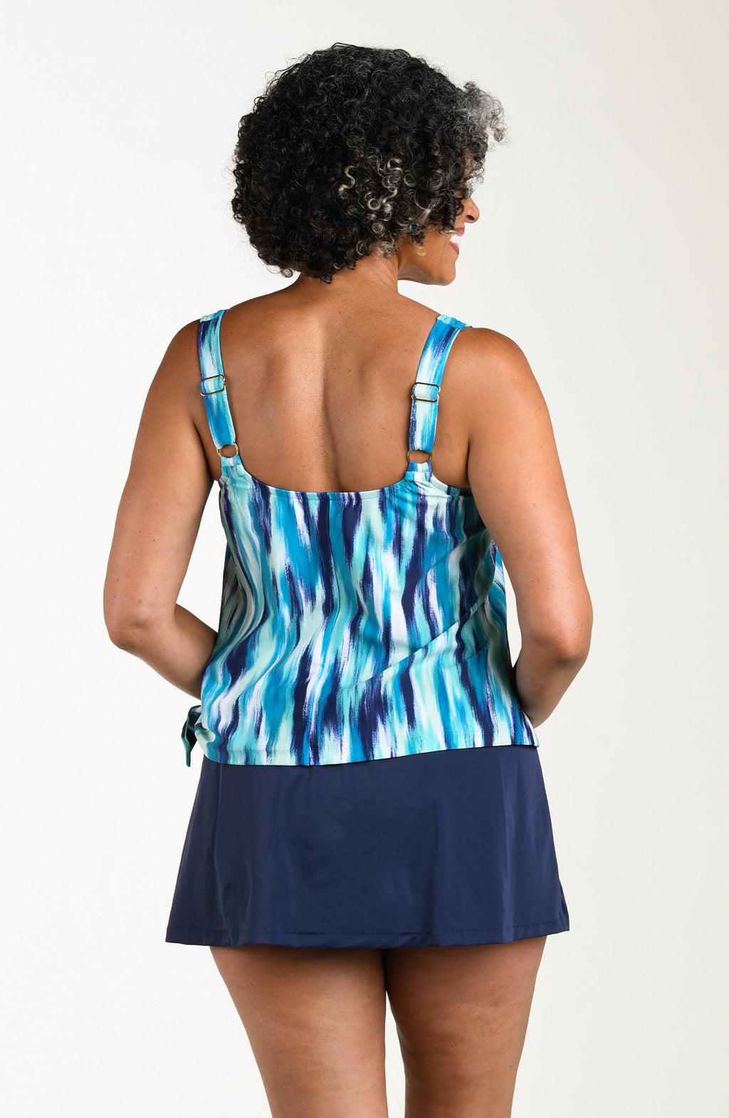 Back image of model wearing a multicolored teal colored one-piece featuring a scoop neckline, over the shoulder straps, and side tie detail. The top looks like a tankini and the bottom is solid indigo skirt.