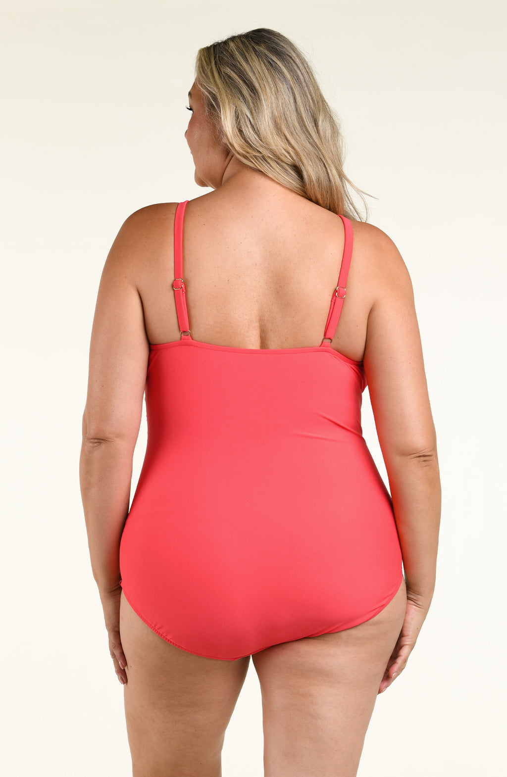 A woman modeling a coral red halter-neck one-piece swimsuit against a plain white studio background.