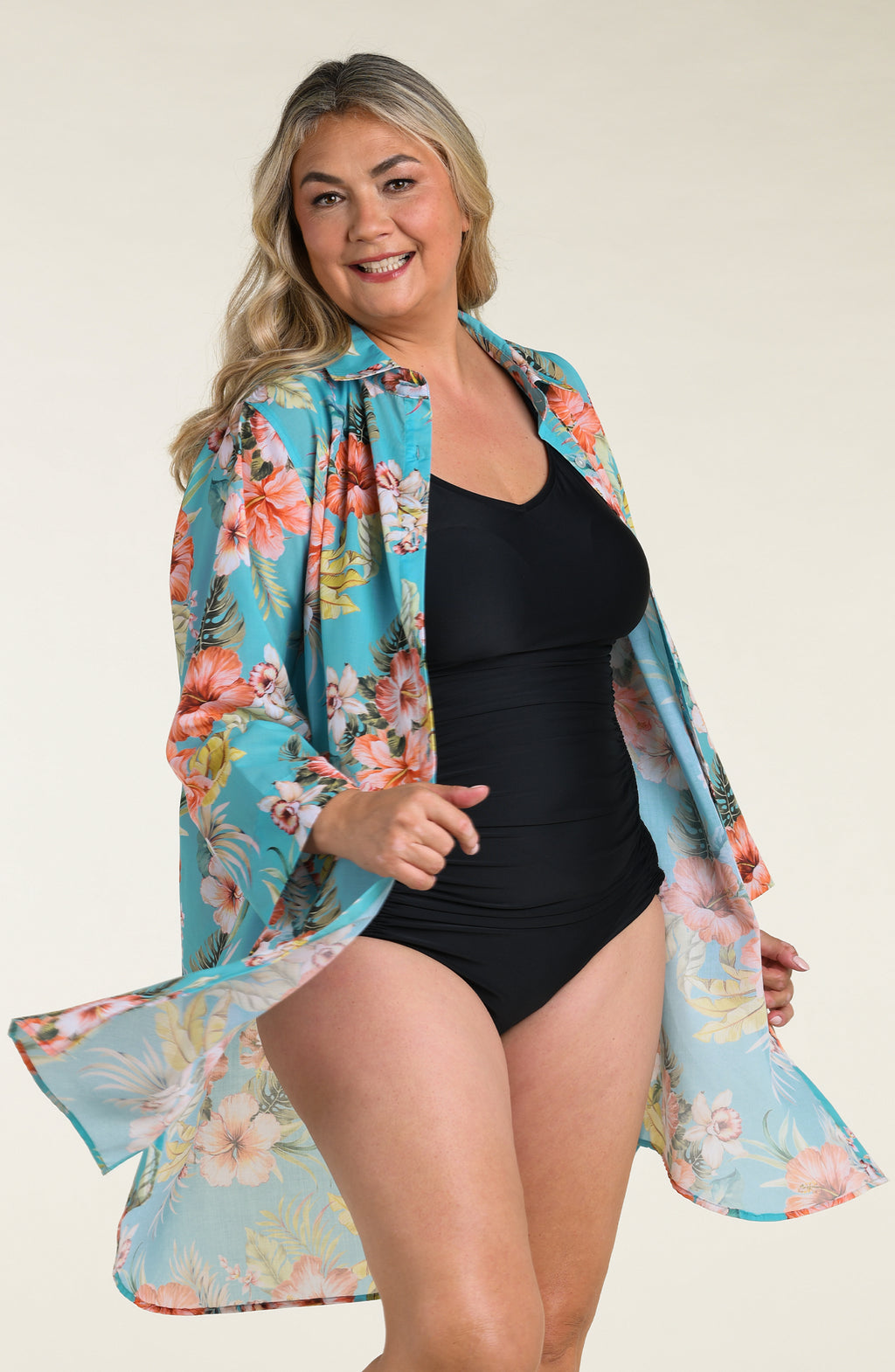 Woman wearing a floral cover up tunic with a black one piece swimsuit underneath, against a white background