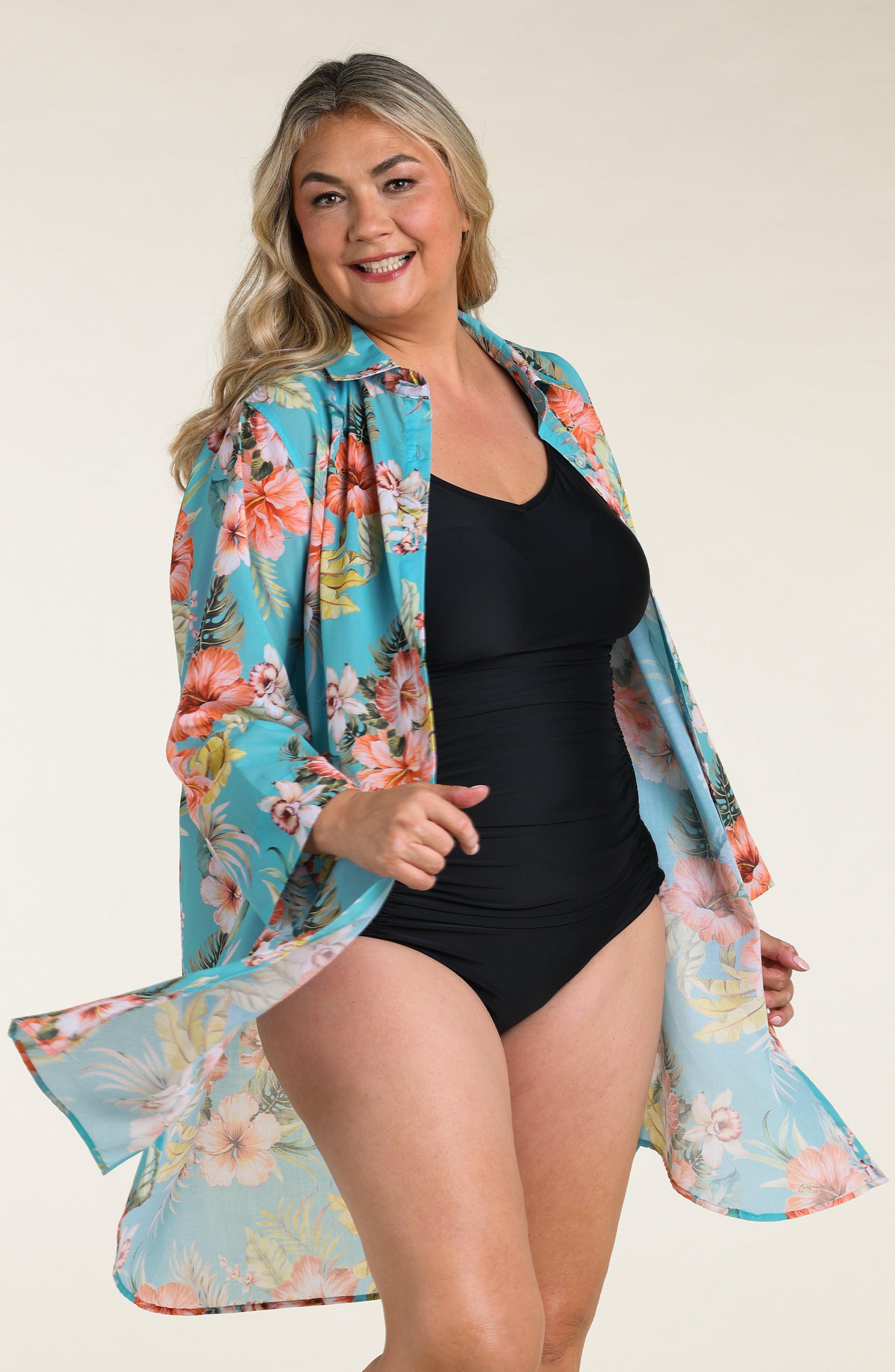 Woman wearing a floral cover up tunic with a black one piece swimsuit underneath, against a white background