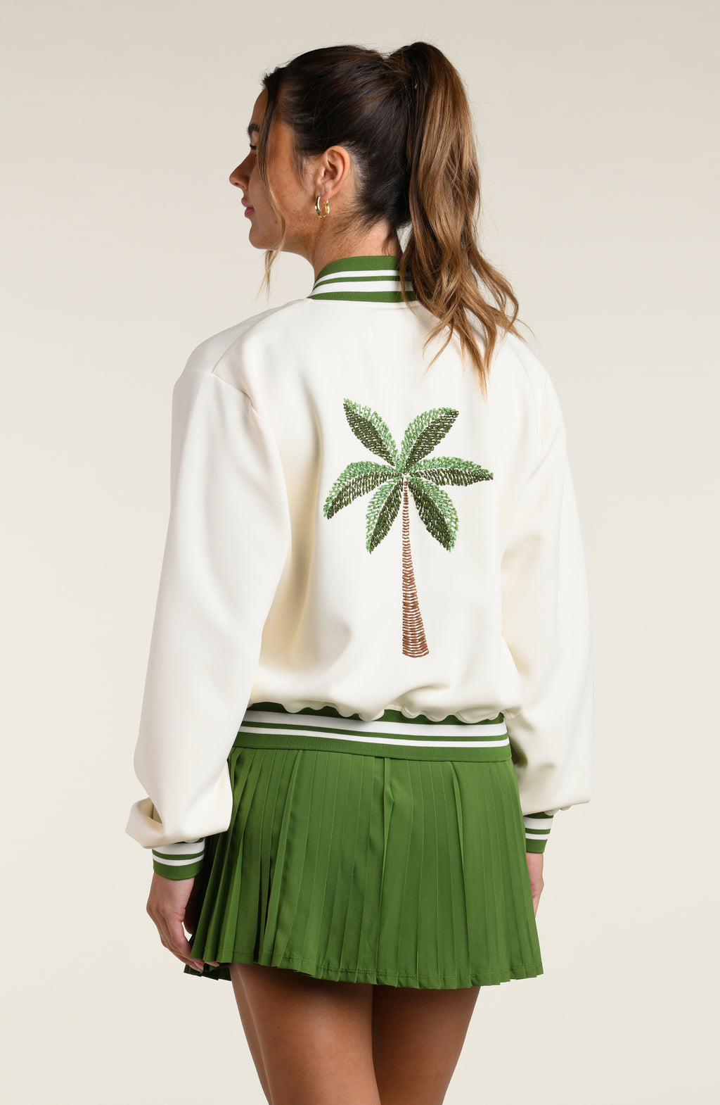 Woman wearing a green sports bra, matching skort and white jacket with a palm tree logo, against a white background