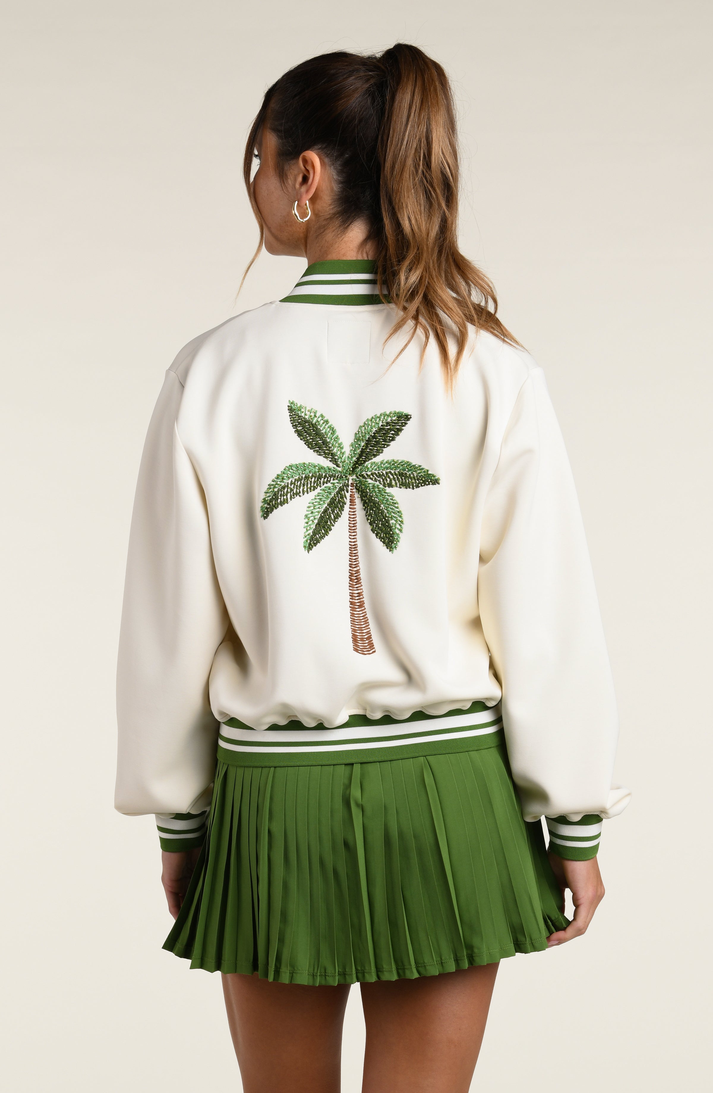 Woman wearing a green sports bra, matching skort and white jacket with a palm tree logo, against a white background