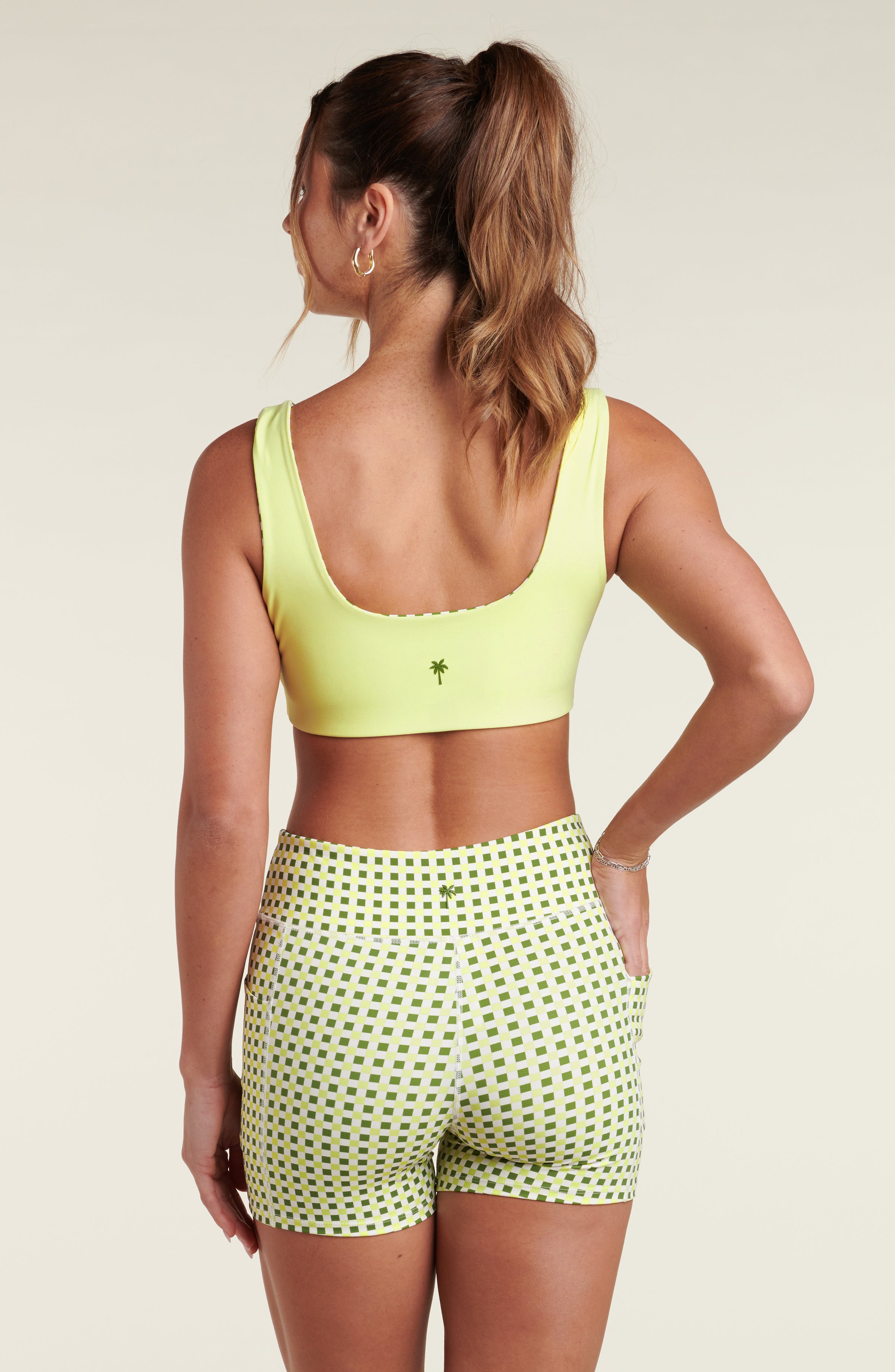 Woman wearing a lime green sports bra and green and white checkered bike shorts against a white background.