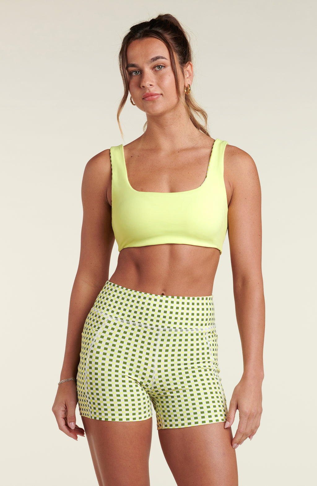 Woman wearing a lime green sports bra and green and white checkered bike shorts against a white background.