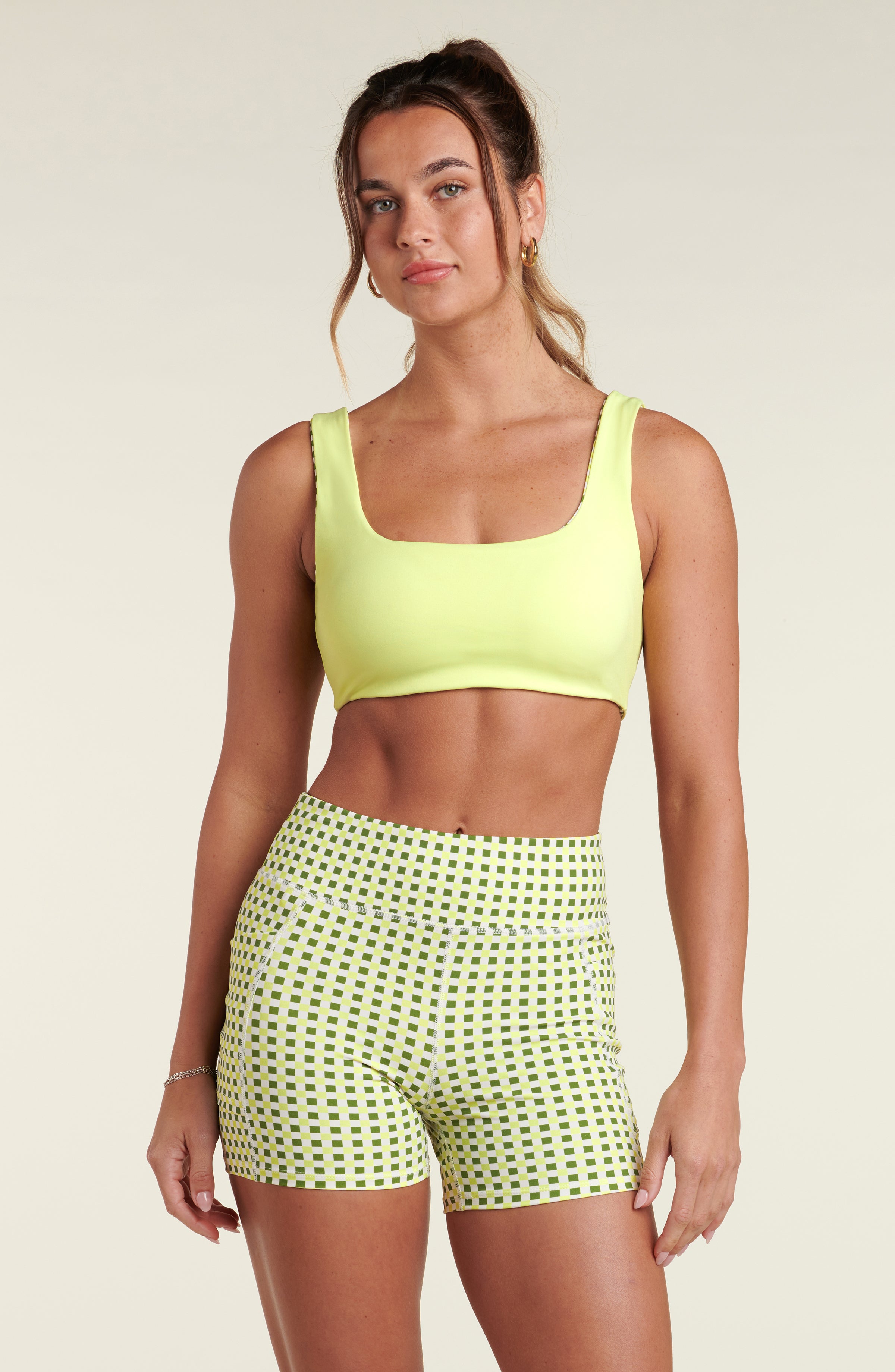 Woman wearing a lime green sports bra and green and white checkered bike shorts against a white background.