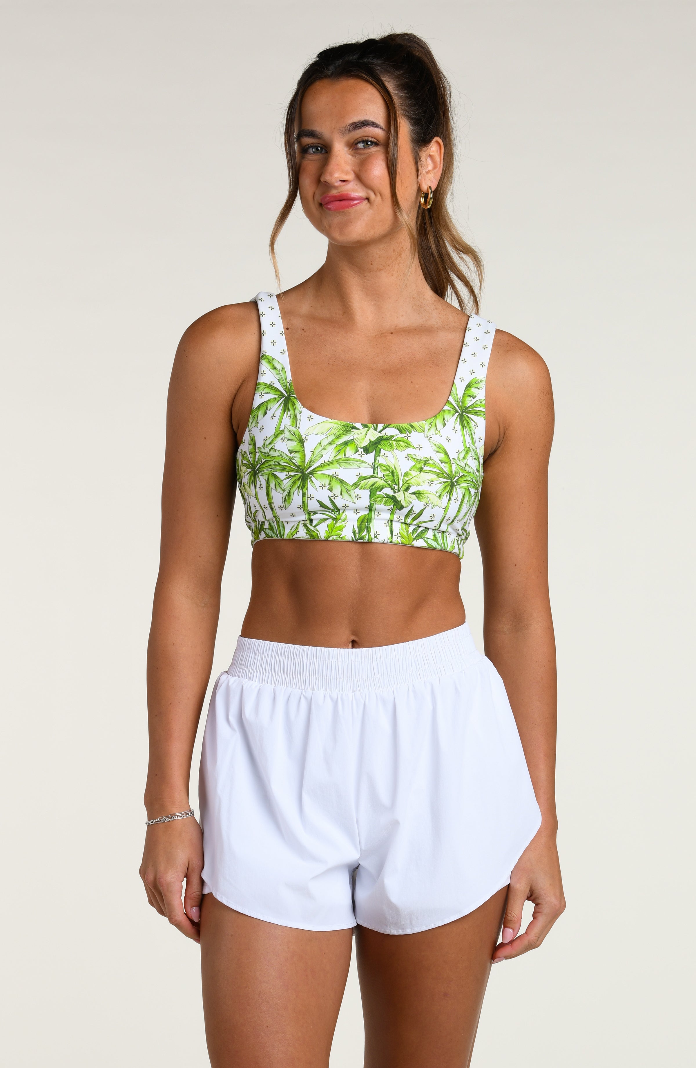 Woman wearing a white sports bra with green palm tree pattern and a white swim skort, worn by a person against a white background