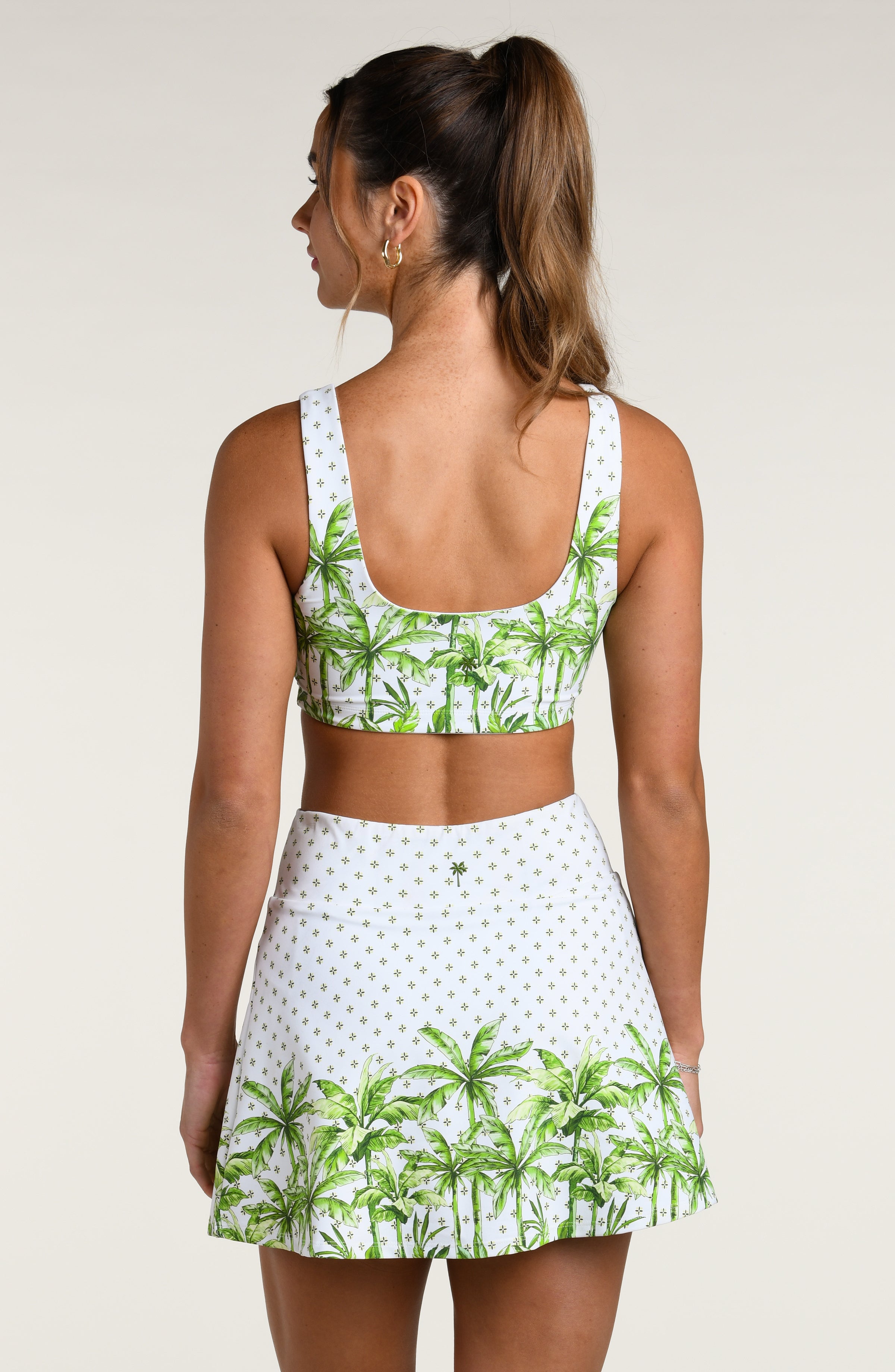 Woman wearing a white sports bra with green palm tree pattern and matching swim skort, worn by a person against a white background