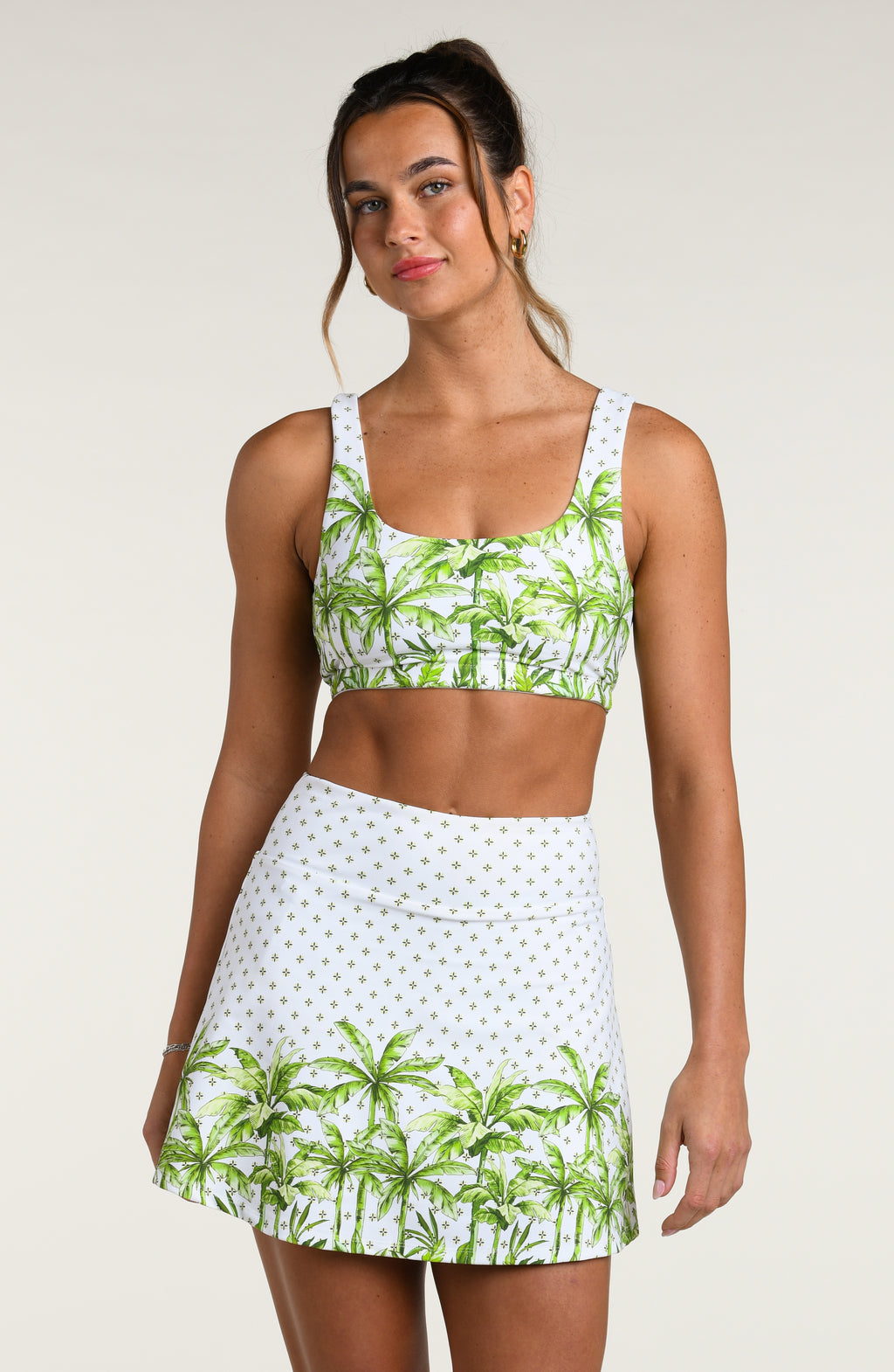 Woman wearing a white sports bra with green palm tree pattern and matching swim skort, worn by a person against a white background