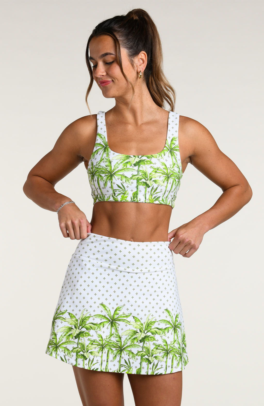 Woman wearing a white sports bra with green palm trees and a matching skort against a white background. 