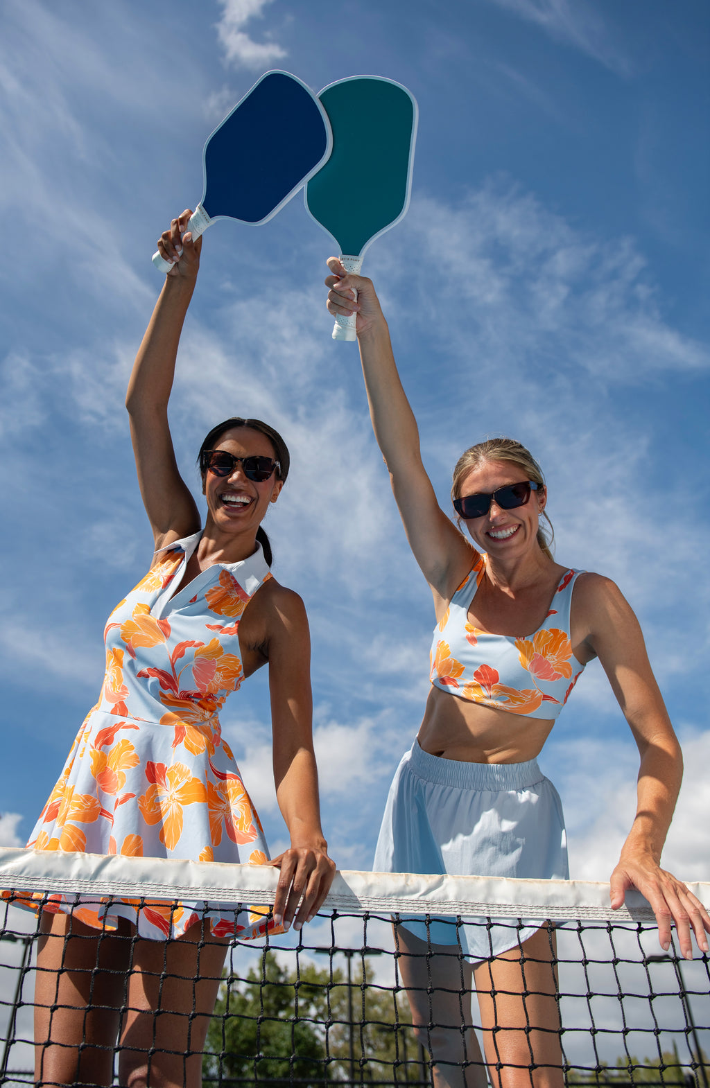 Two women on a pickleball court holding up blue and green paddles against a blue sky.