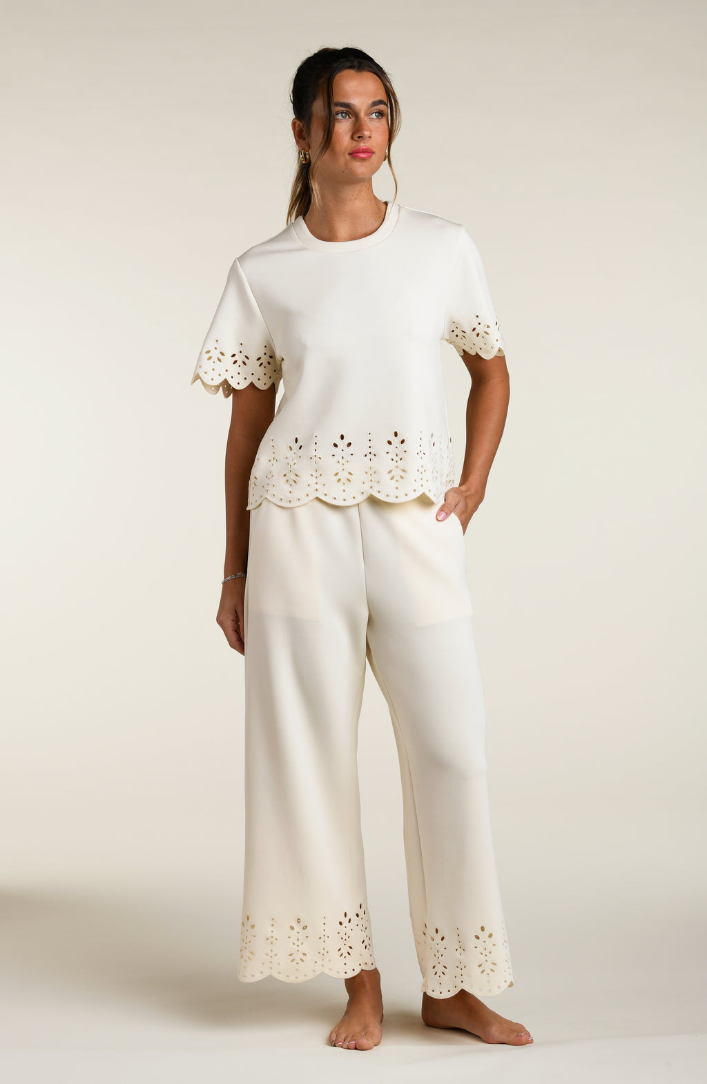 Woman wearing a beige short sleeve top with scalloped edges and a matching pants on a plain background