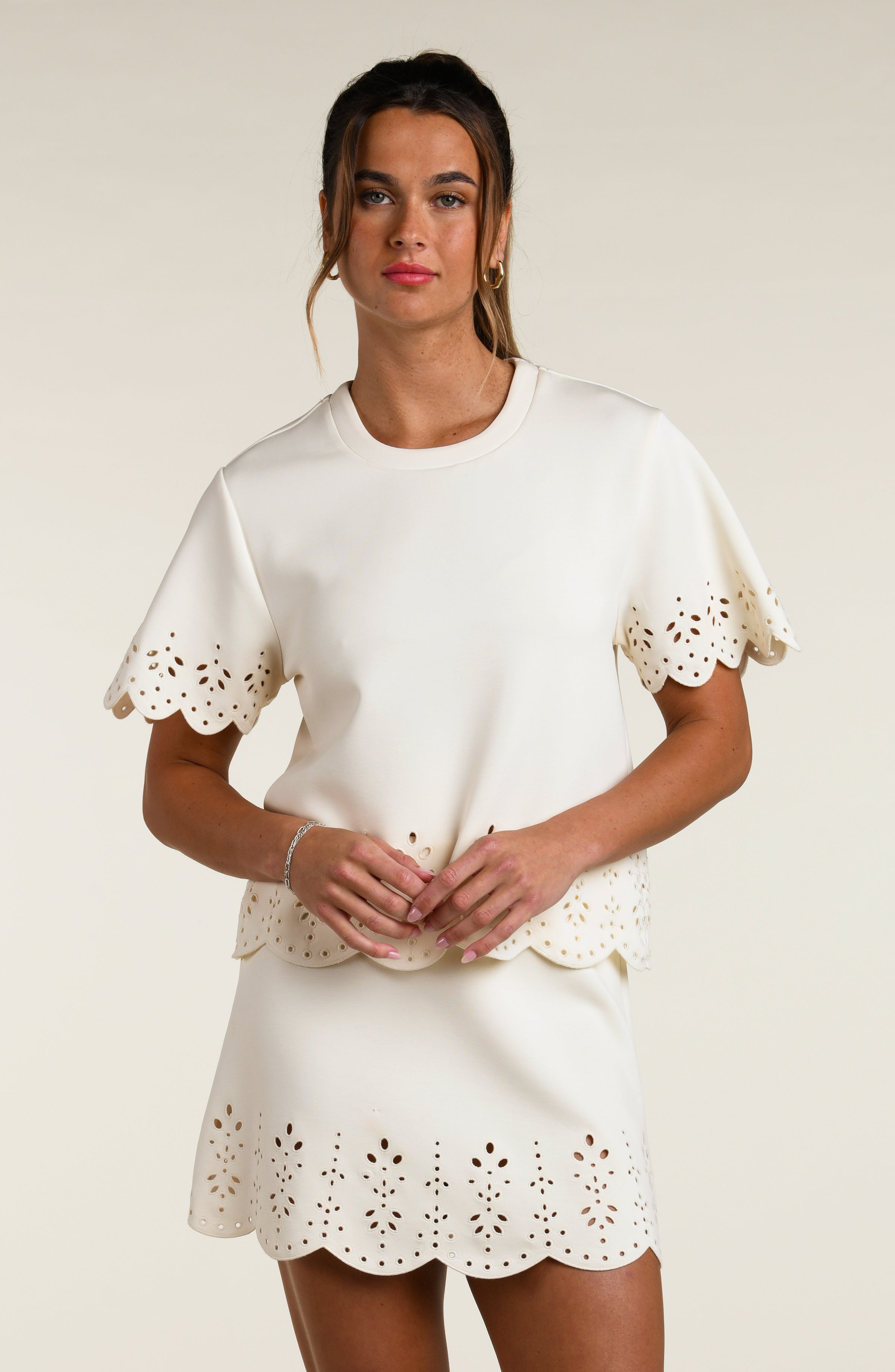 Woman wearing a beige short sleeve top with scalloped edges and a matching skirt on a plain background