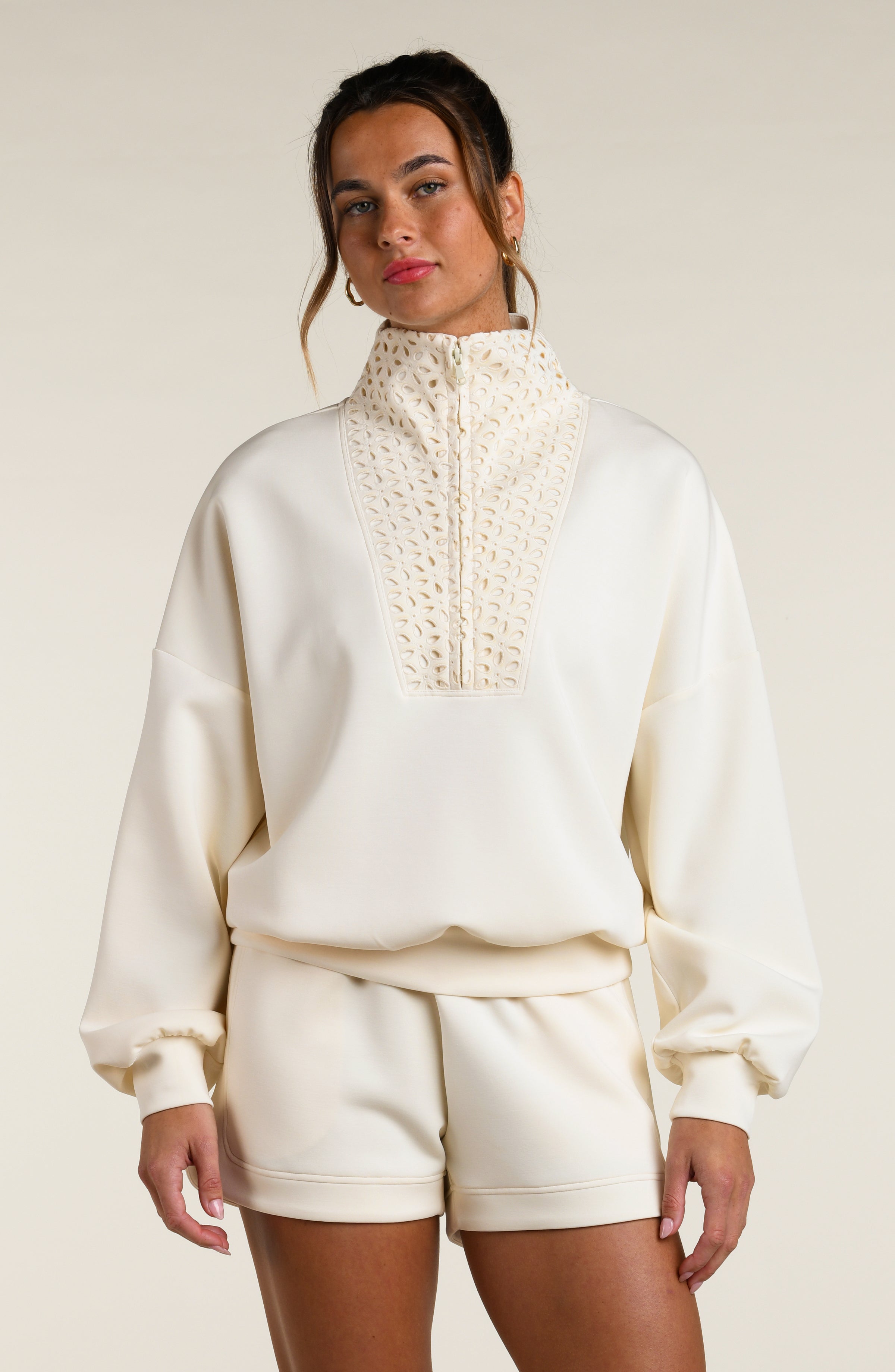 Woman wearing a cream-colored sweatshirt with a high-neck top and shorts on a plain background