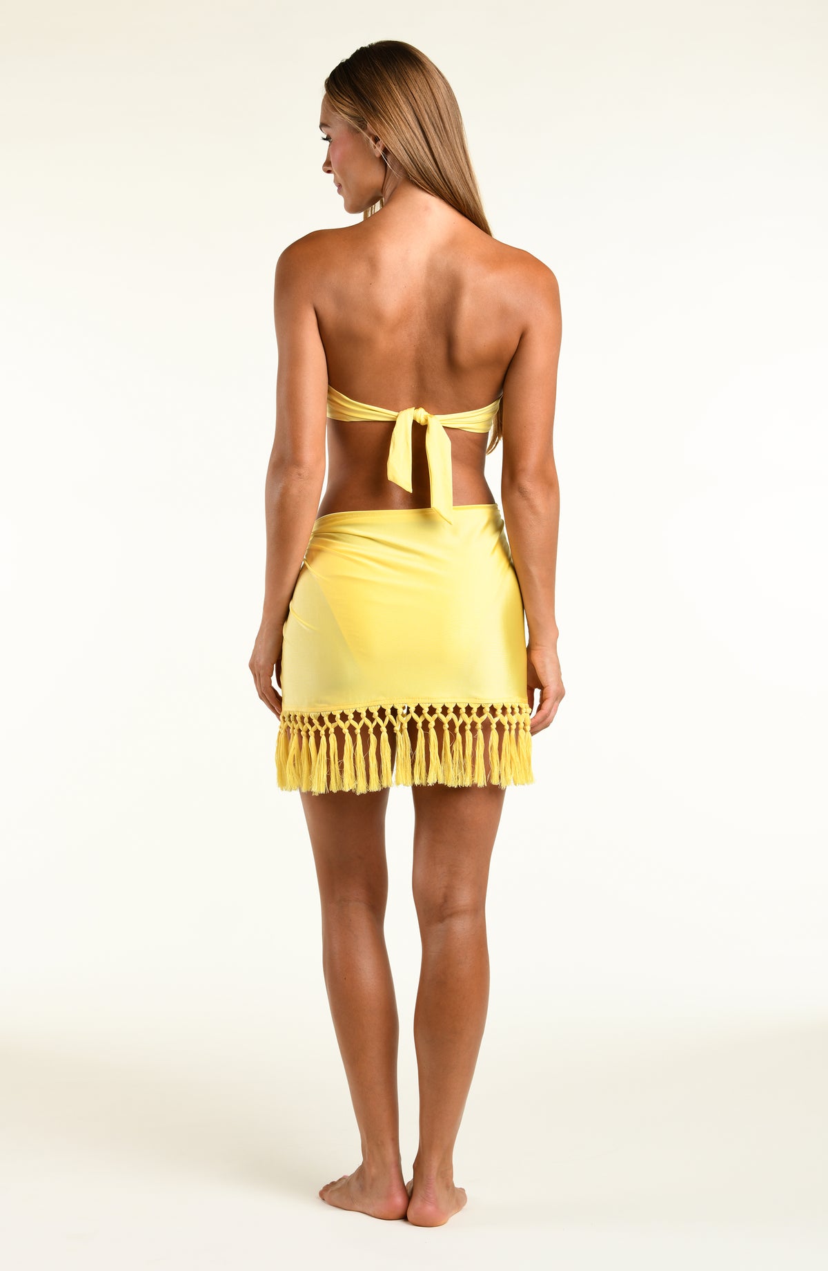 Woman wearing a bright yellow bandeau bikini top and short sarong against a white background. 