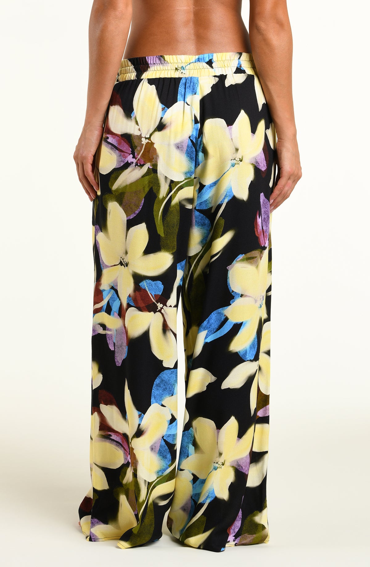 A model wearing wide-leg black pants with a vibrant yellow, blue, and purple floral print, standing against a plain white studio background.
