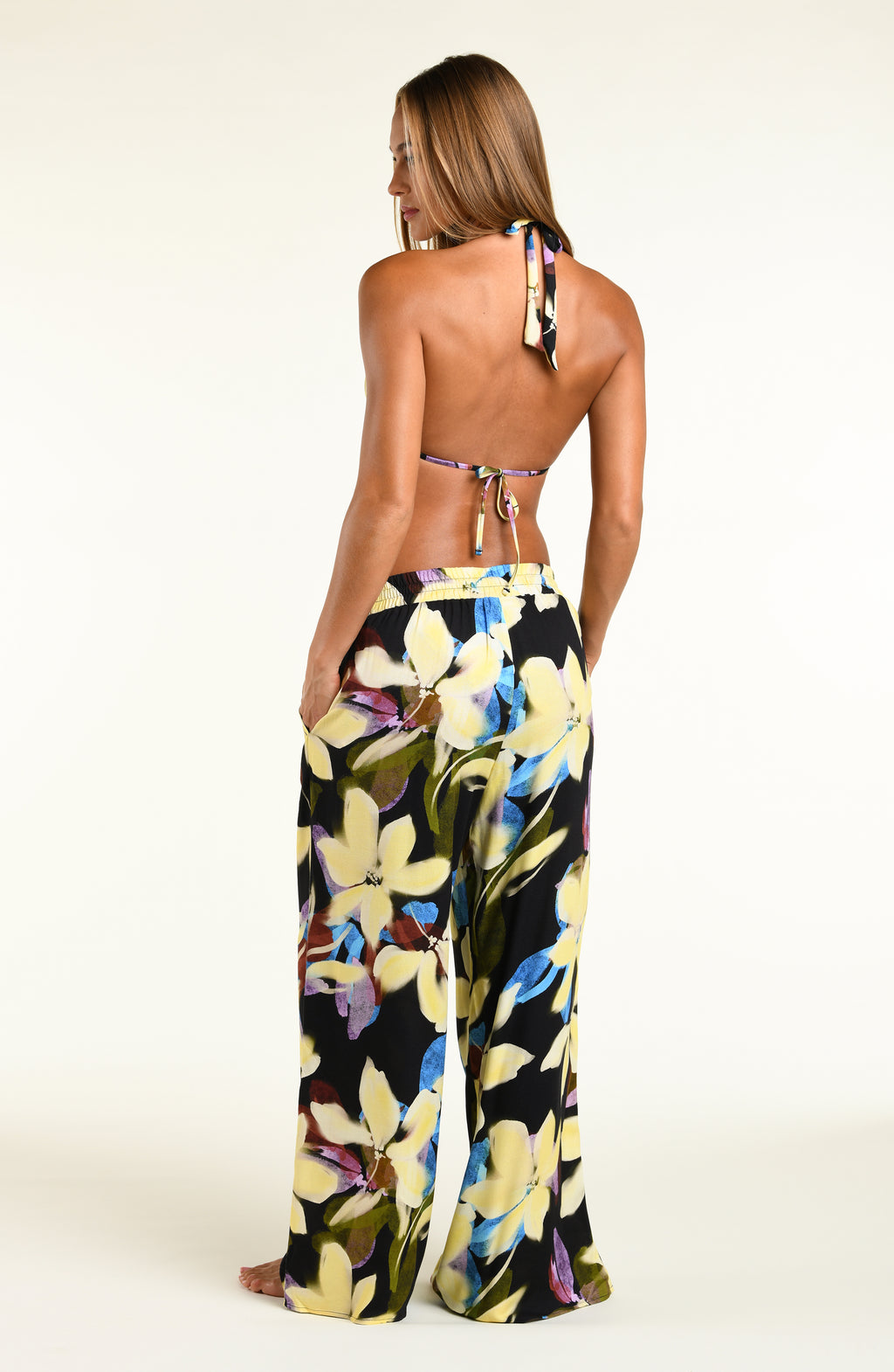 A model wearing a bikini top and matching beach pants with a vibrant yellow, blue, and purple floral print, standing against a plain white studio background.
