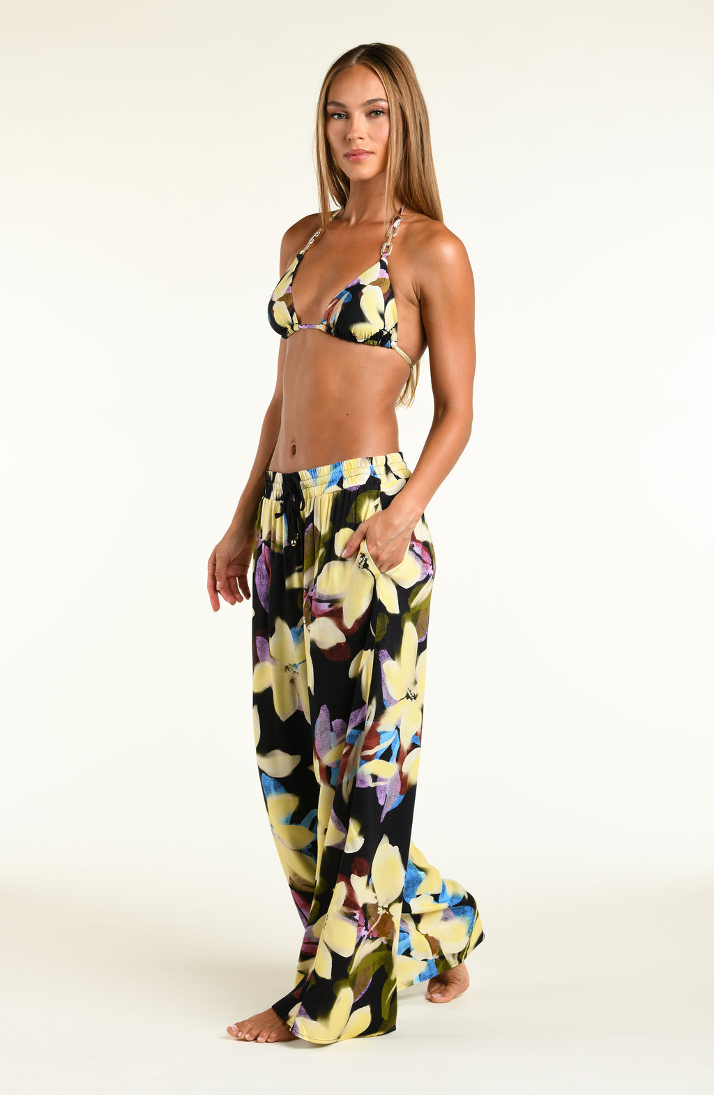 A model wearing a bikini top and matching beach pants with a vibrant yellow, blue, and purple floral print, standing against a plain white studio background.