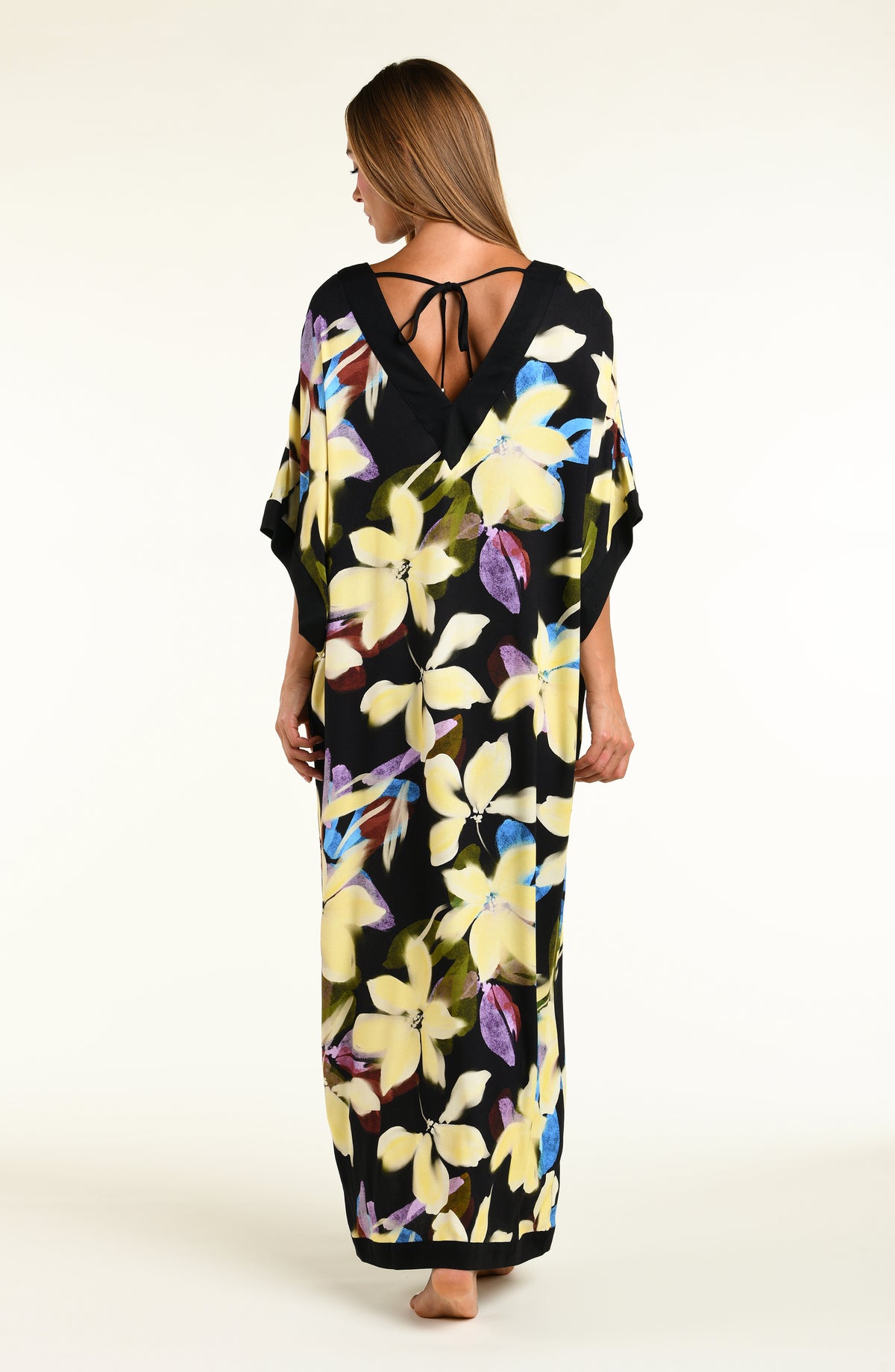 A woman modeling a black maxi caftan with a large yellow, green, and blue floral print, set against a plain off-white studio background.