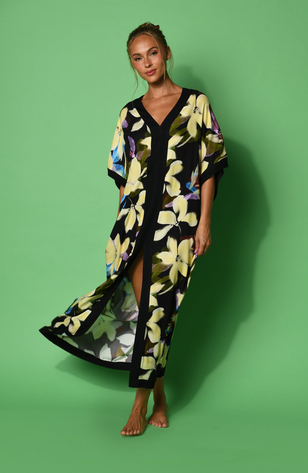 A woman modeling a black maxi caftan with a large yellow, green, and blue floral print, set against a green background.