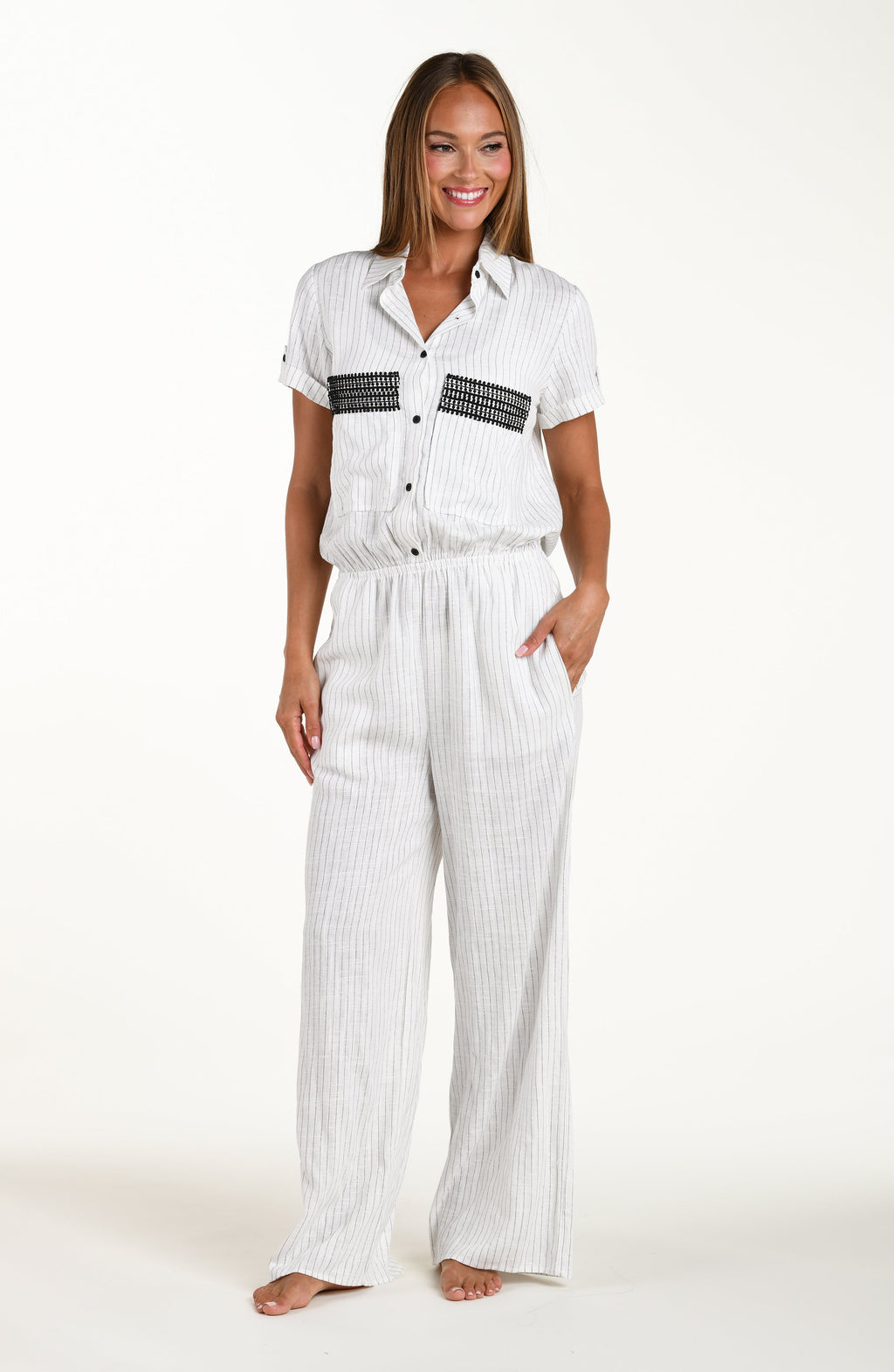 Full body front image of model wearing a pant jumpsuit in a button up collared front with wide pockets adorned with crochet trim, short sleeves, and pant pockets. Featuring a cream colored linen print. 