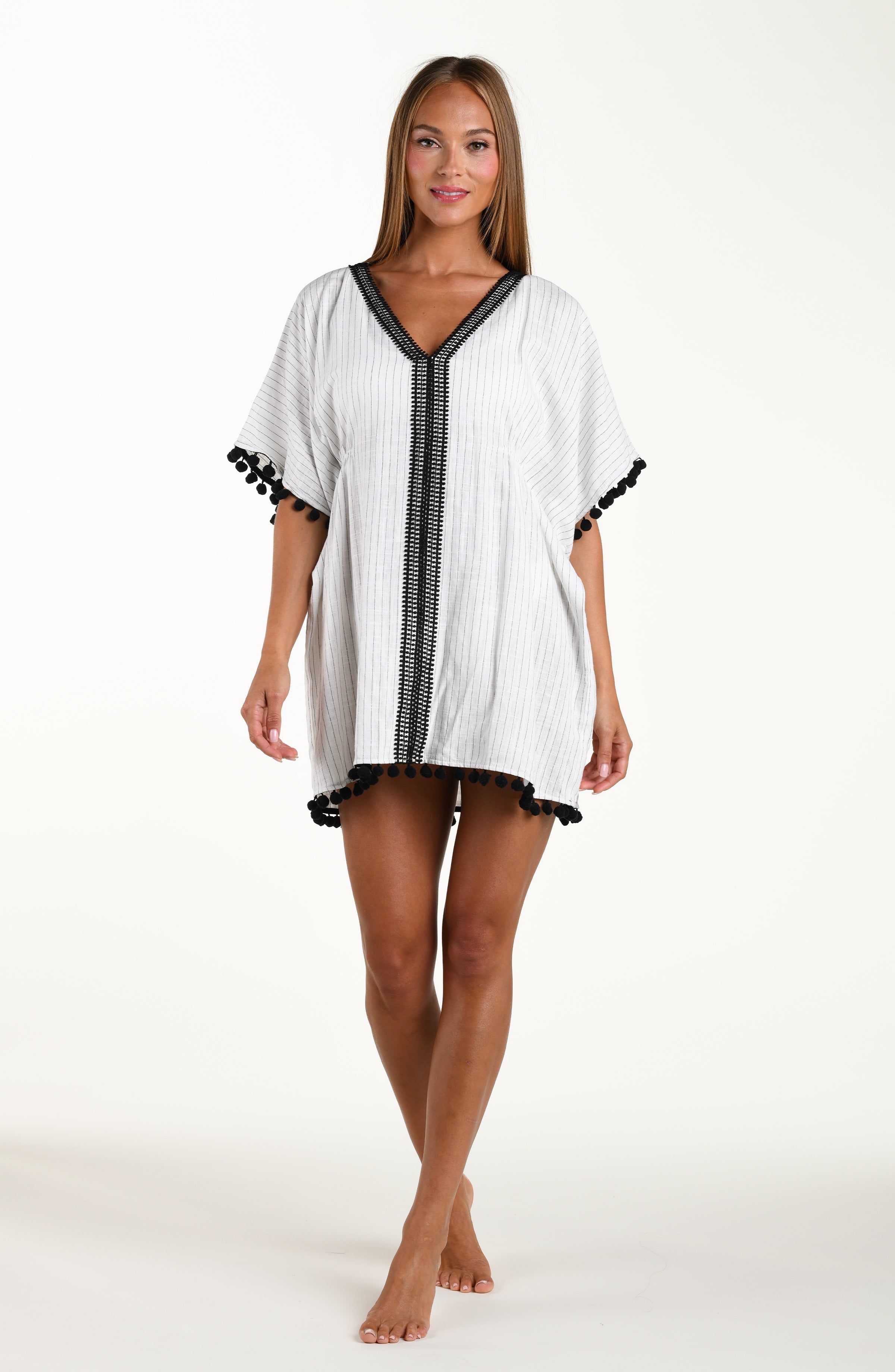 Full body front image of model wearing a short pull over v-neck caftan with three quarter sleeves adorned with tassel trim. Featuring a cream colored linen print.