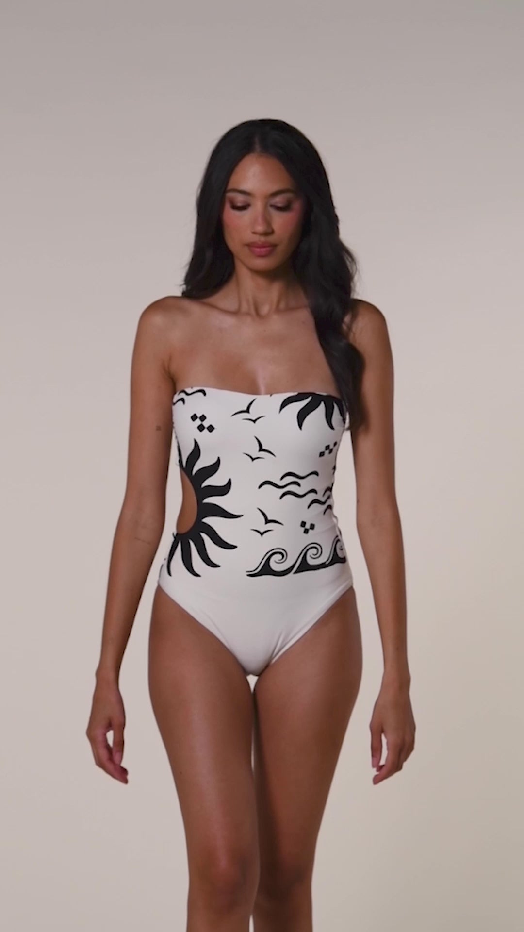 Video of a woman wearing a white one piece bandeau swimsuit with black designs on a beige background. 