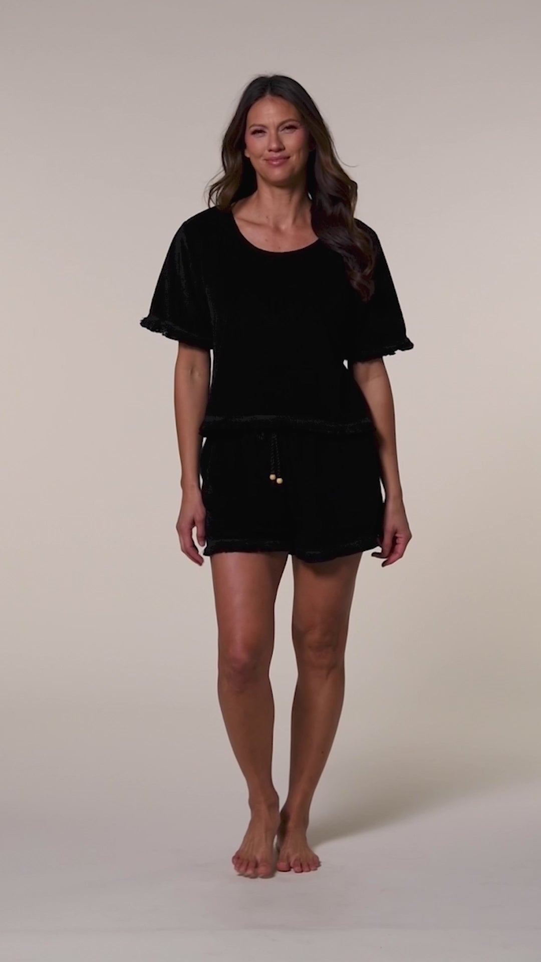 Video of a woman wearing a black short sleeve top and matching beach shorts against a beige background.