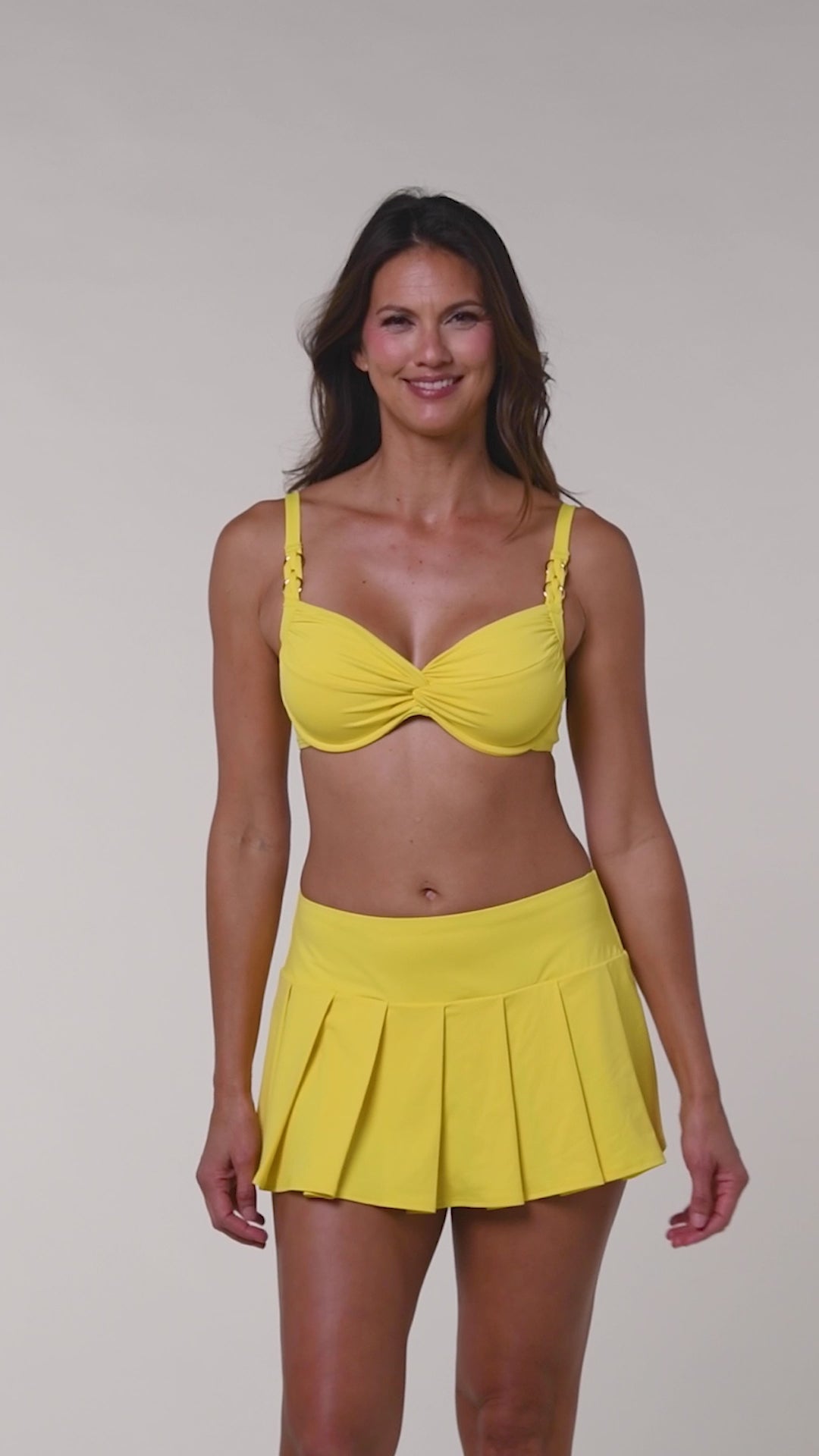 Video of a woman wearing a yellow bikini top and a matching swim skirt bottom against a beige background.