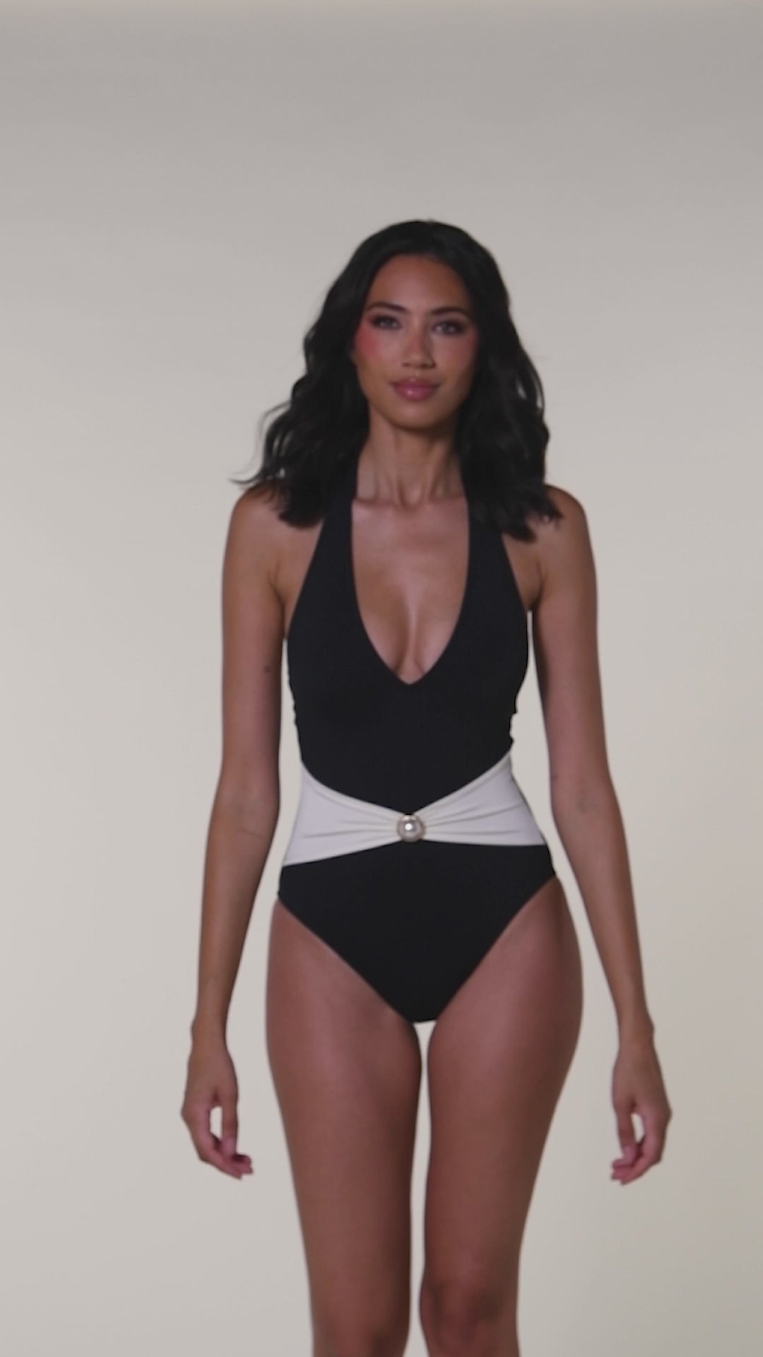 Video of a woman modeling a one-piece swimsuit with a black color-blocked design, featuring a white sash at the waist with a small pearl detail, set against a white background.