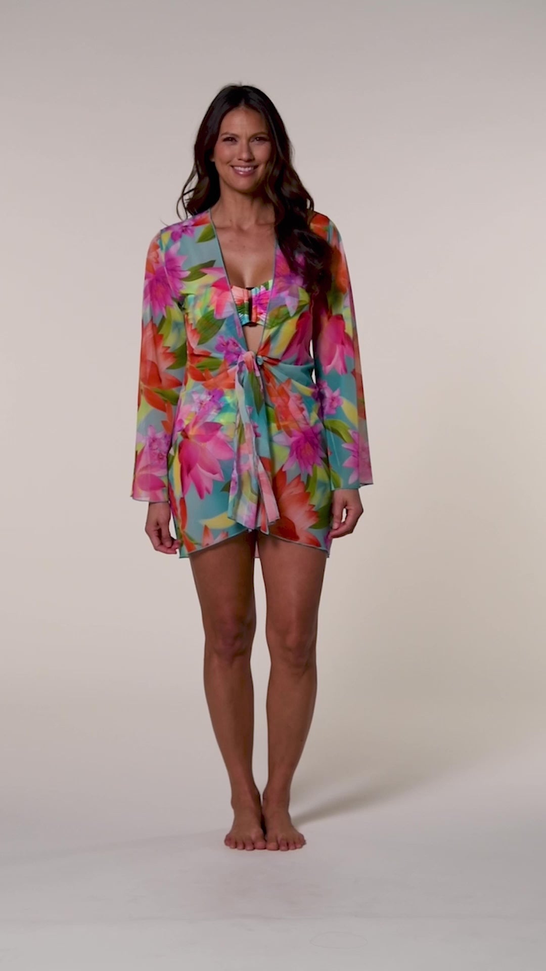 Video of a woman wearing a bright floral kimono cover up with a matching bikini underneath, against a beige background.