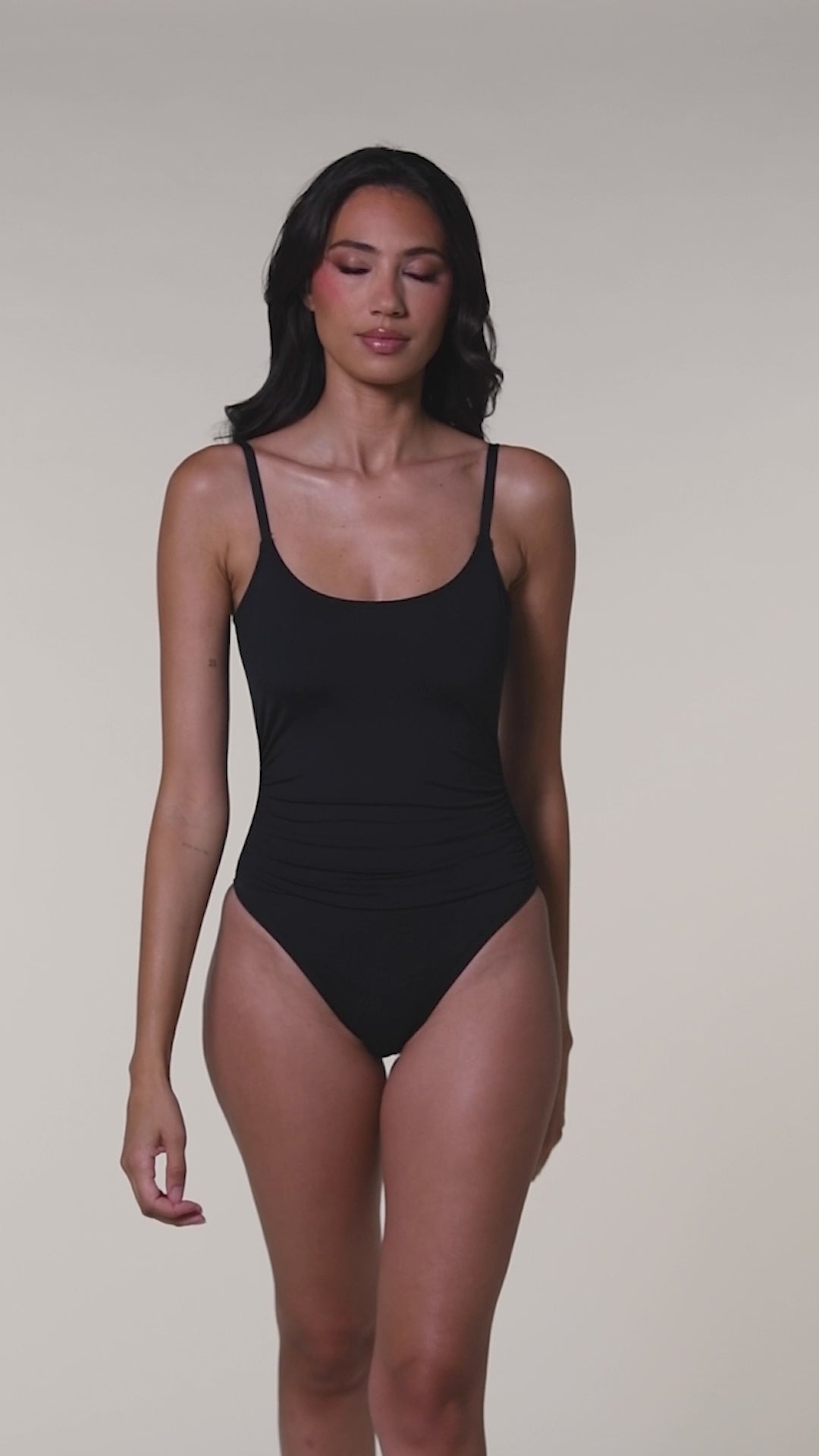 Video of a woman wearing a solid black one piece swimsuit with a scoop neckline and over the shoulder straps against a white background. 