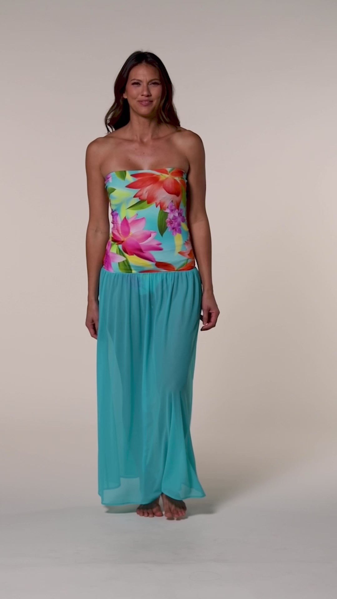 Video of a woman wearing a turquoise and bright floral convertible maxi dress cover up against a beige background. 
